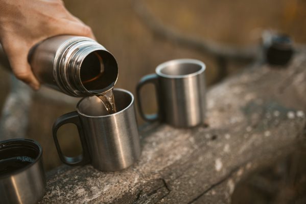 Male hand is pouring a hot drink from a thermos into metal camping cups standing on a fallen tree. Time for a break in nature. Close up shot.