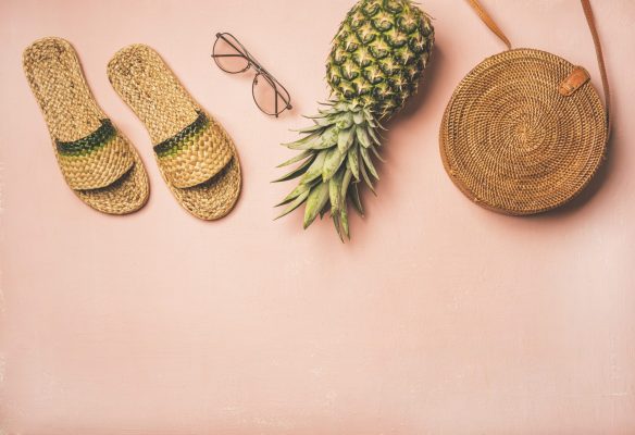 Variety of summer apparel items. Flat-lay of summer flip flops, sunglasses, round wicker shoulder bag and pineapple over pastel pink background, top view, copy space. Summer fashion garment concept
